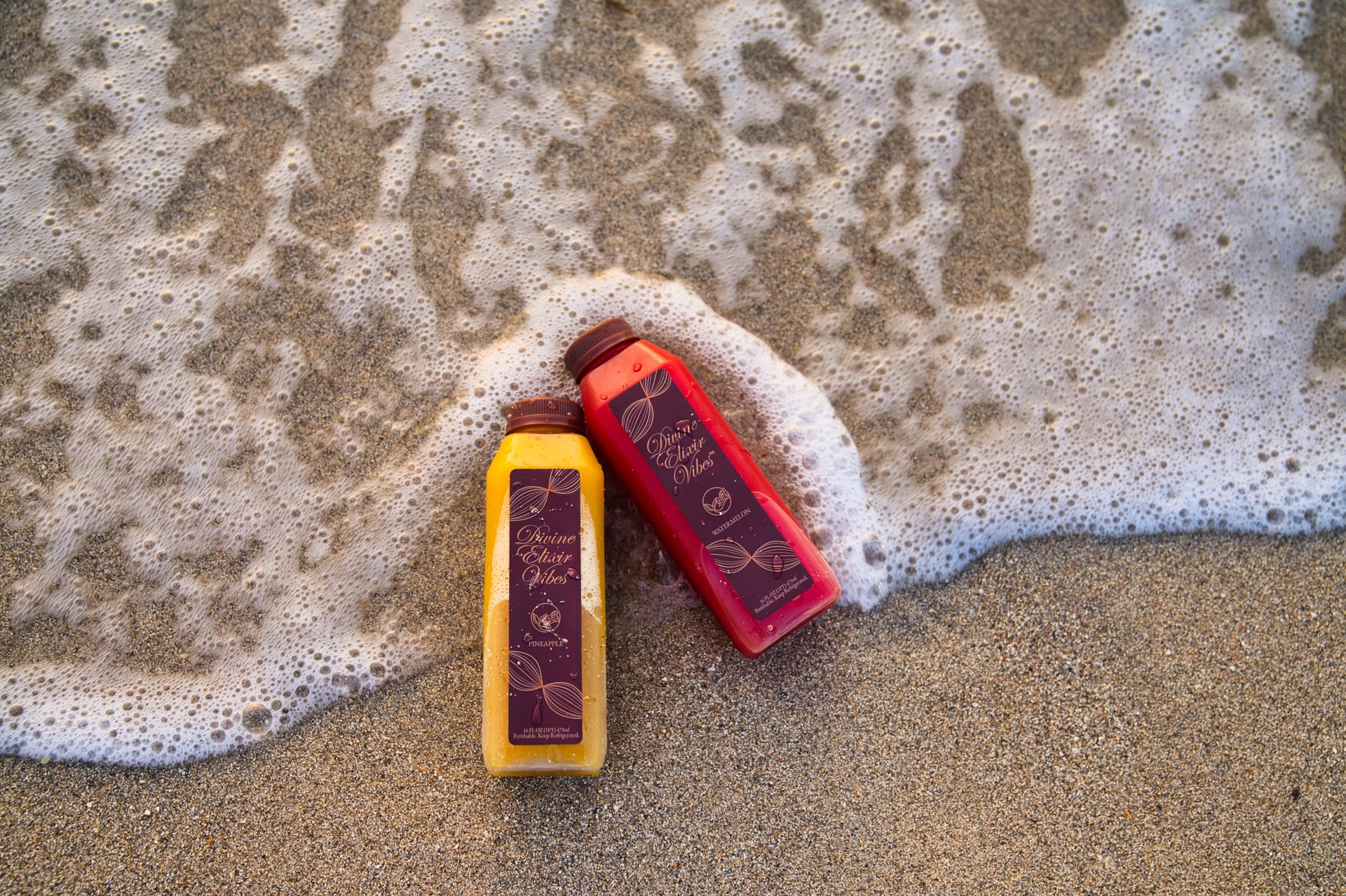 Divine Elixir Vibes pineapple and watermelon bottles in sea foam on sand